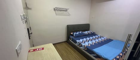 Habitación pequeña y funcional con una cama individual, una mesita, un banco, aire acondicionado instalado en la pared y un perchero para toallas.