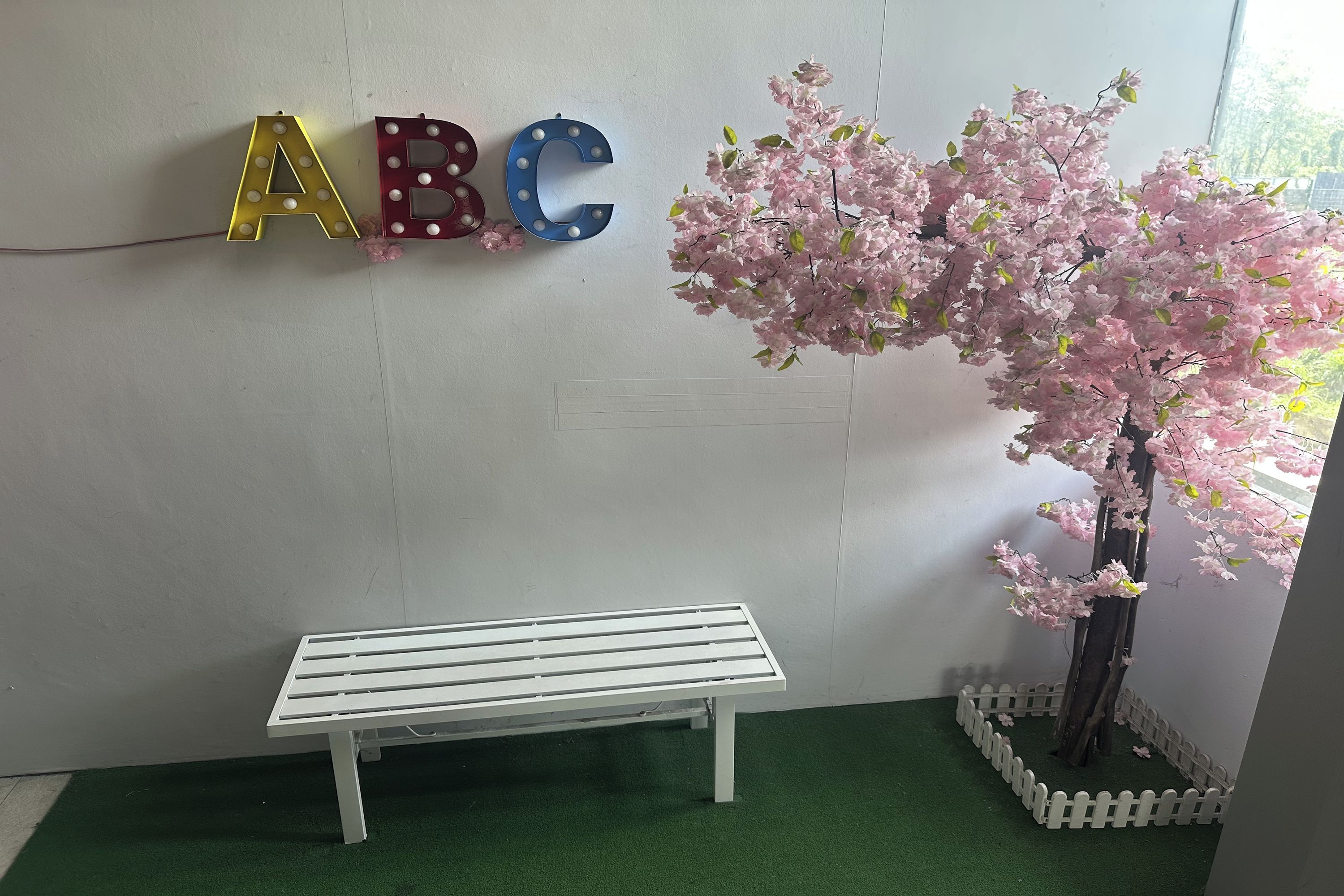 A white bench, a wall with illuminated letters ABC, and a cherry blossom tree.