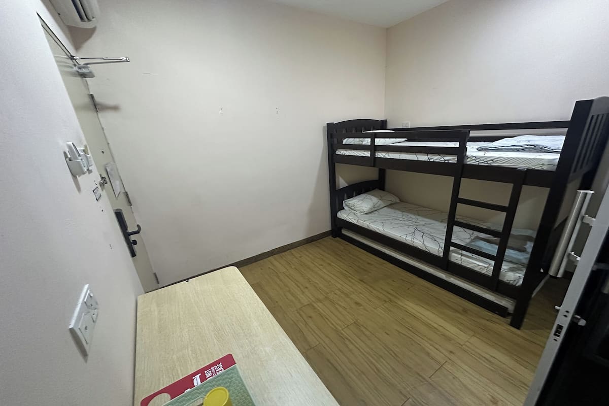 Een kleine, eenvoudig ingerichte kamer met een stapelbed, een houten tafel en een luchtverkoeler aan de muur.