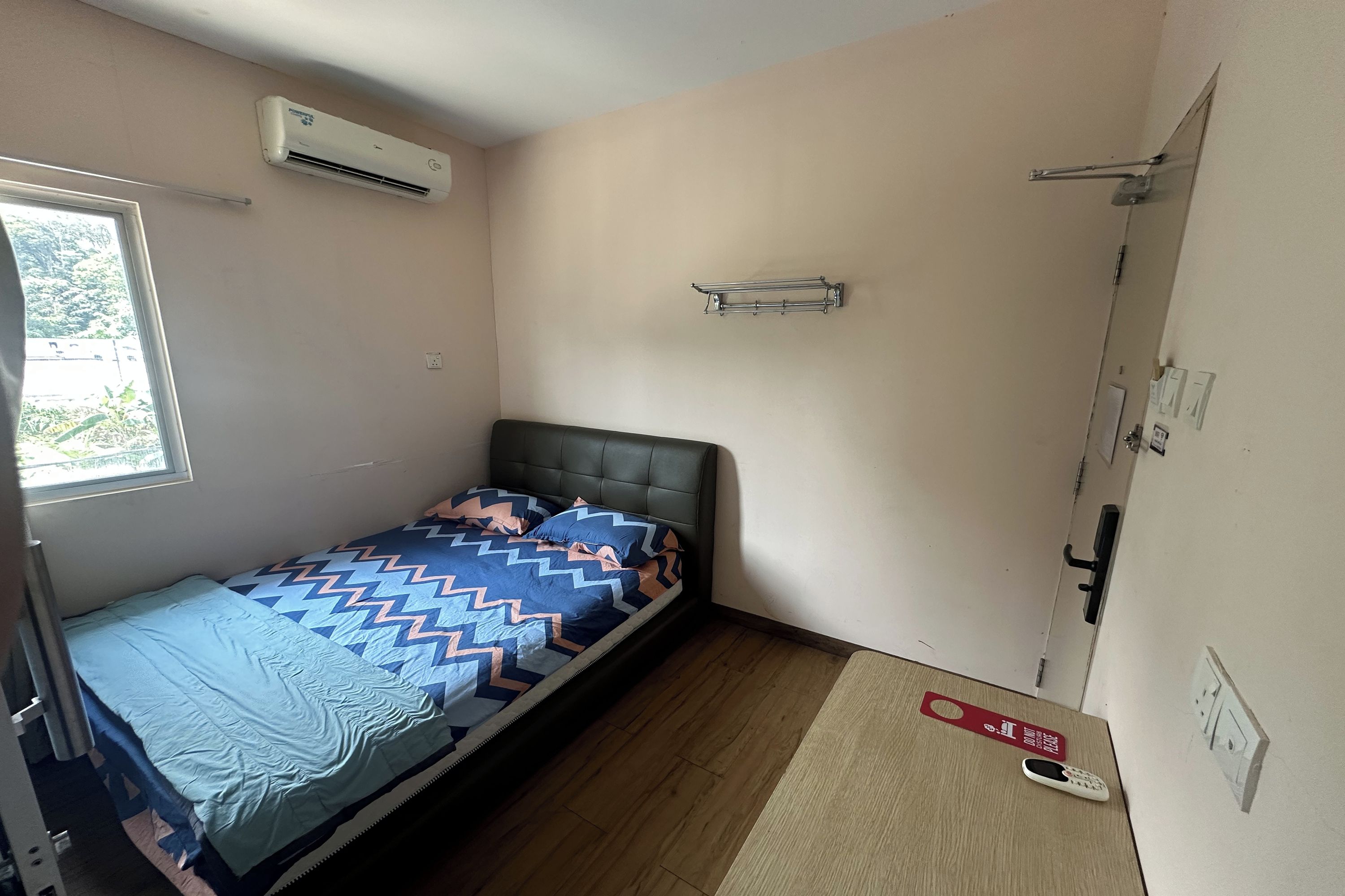 A small, single-bed room with a window, an air conditioner, a bedside table with a remote control, and a door with a handle.
