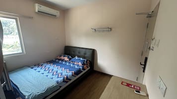 A small, single-bed room with a window, an air conditioner, a bedside table with a remote control, and a door with a handle.