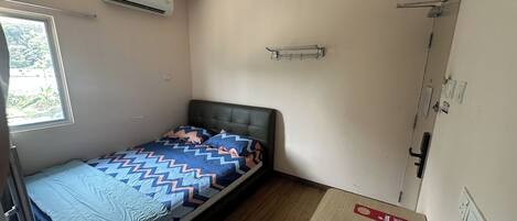 A small, single-bed room with a window, an air conditioner, a bedside table with a remote control, and a door with a handle.