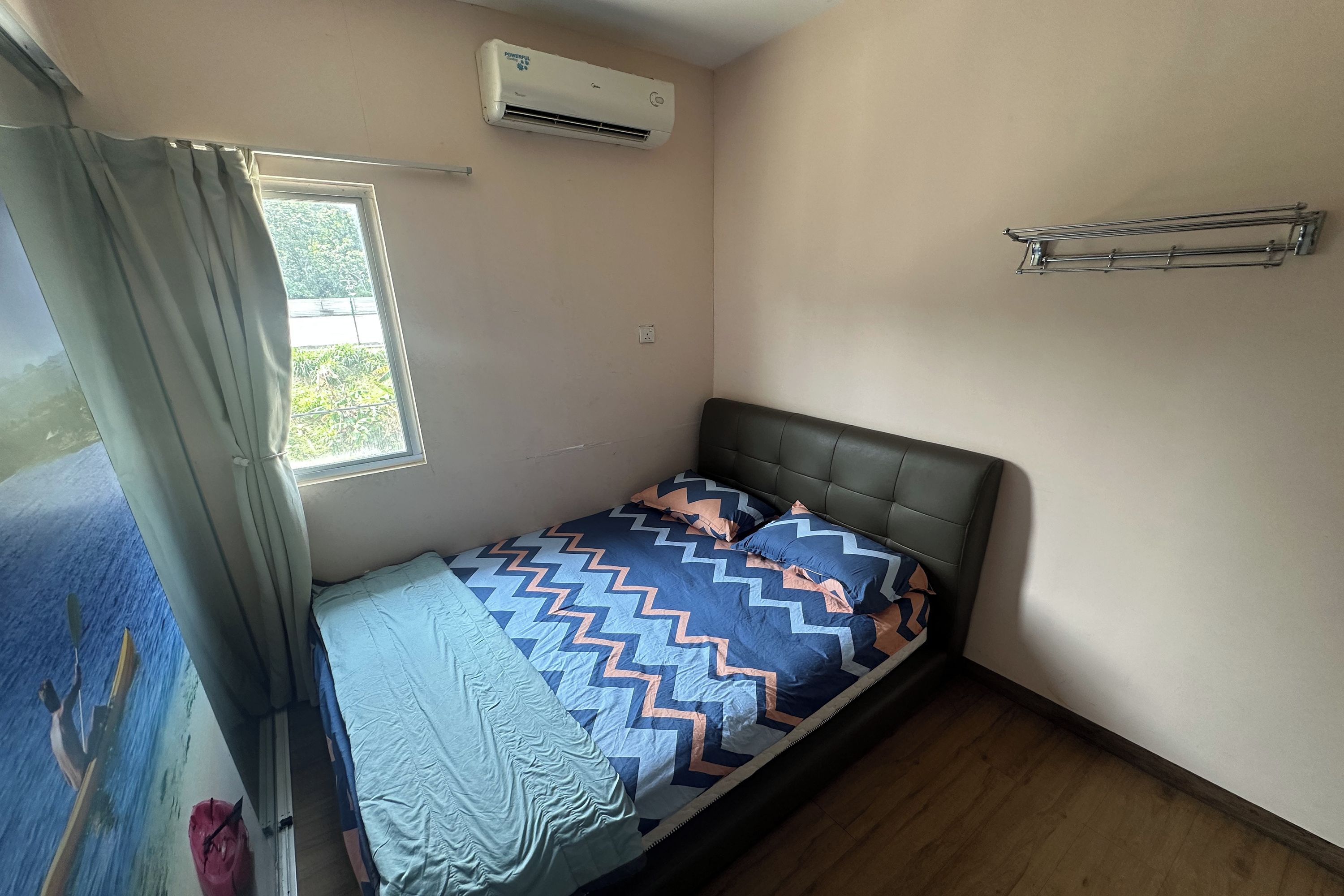 A single bed with a patterned bedspread, a window with curtains, an air conditioning unit, and a metal towel rack.