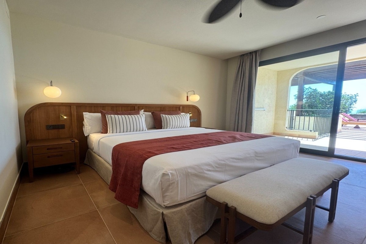 Ein Hotelzimmer mit einem großen Bett, einem Nachttisch, einer Bank und Blick ins Freie.