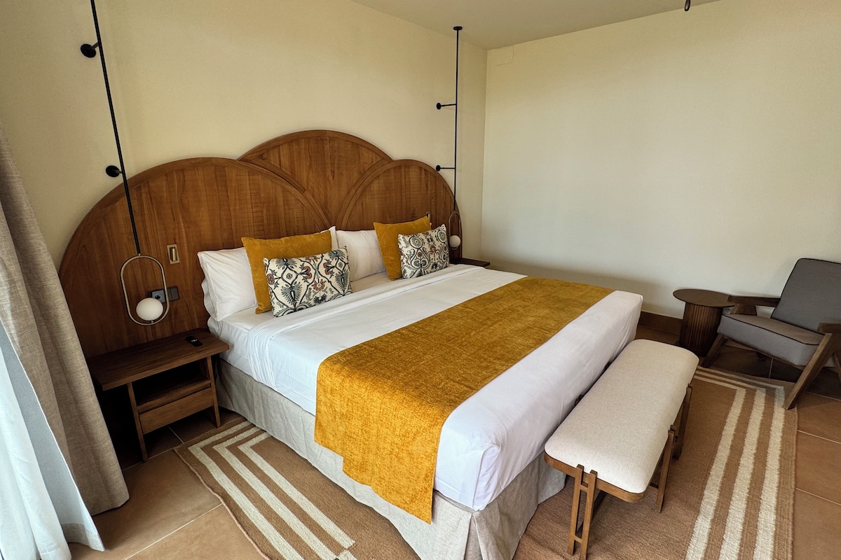 Ein ordentlich eingerichtetes Hotelzimmer mit einem großen Bett, einem Nachttisch, einem Sessel und einer Bank.