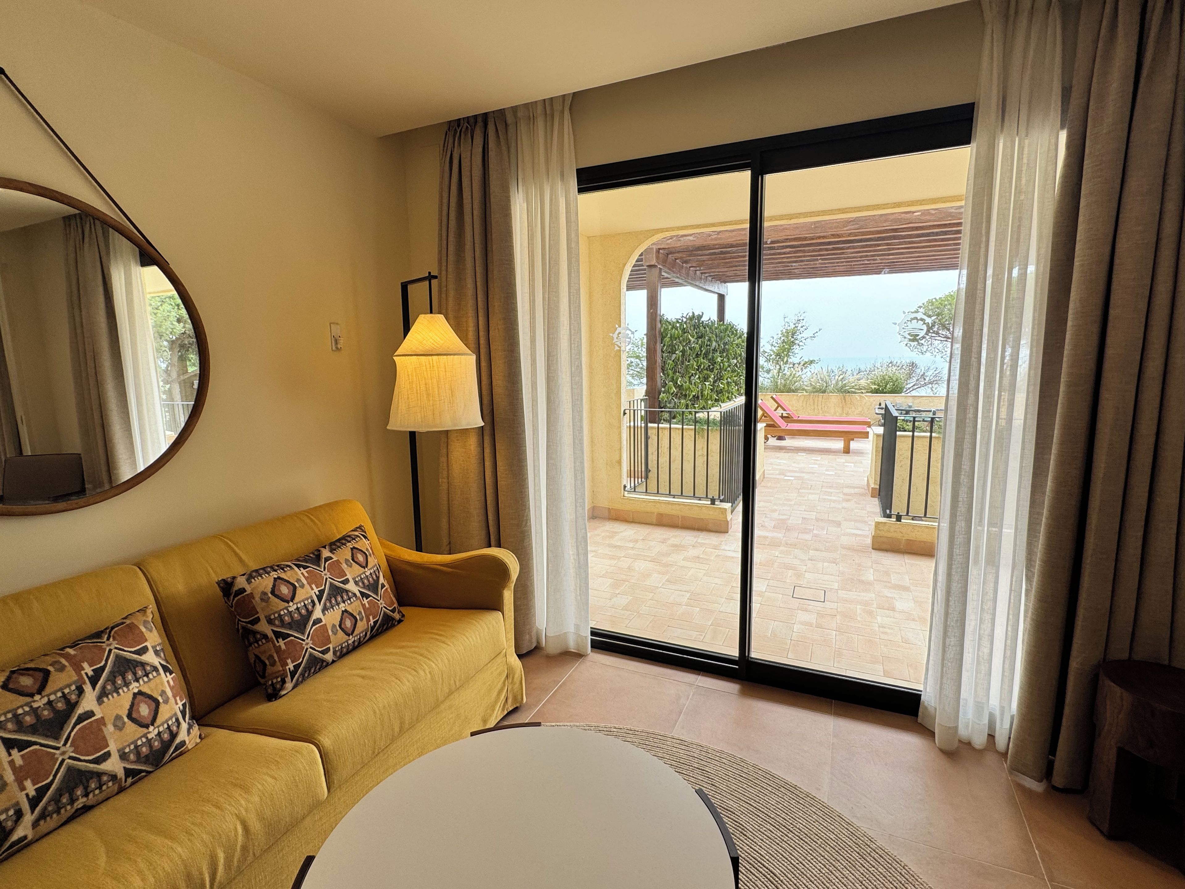 A hotel room with a sofa, a round table, a lamp, and a sliding glass door leading to an outdoor area with a view of the sea.