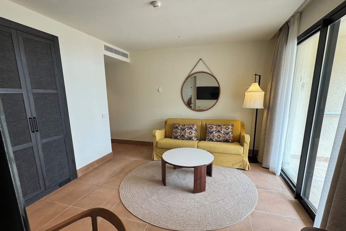 Een woonkamer met een bank, een bijzettafeltje, een stoel, een lamp en een groot raam.