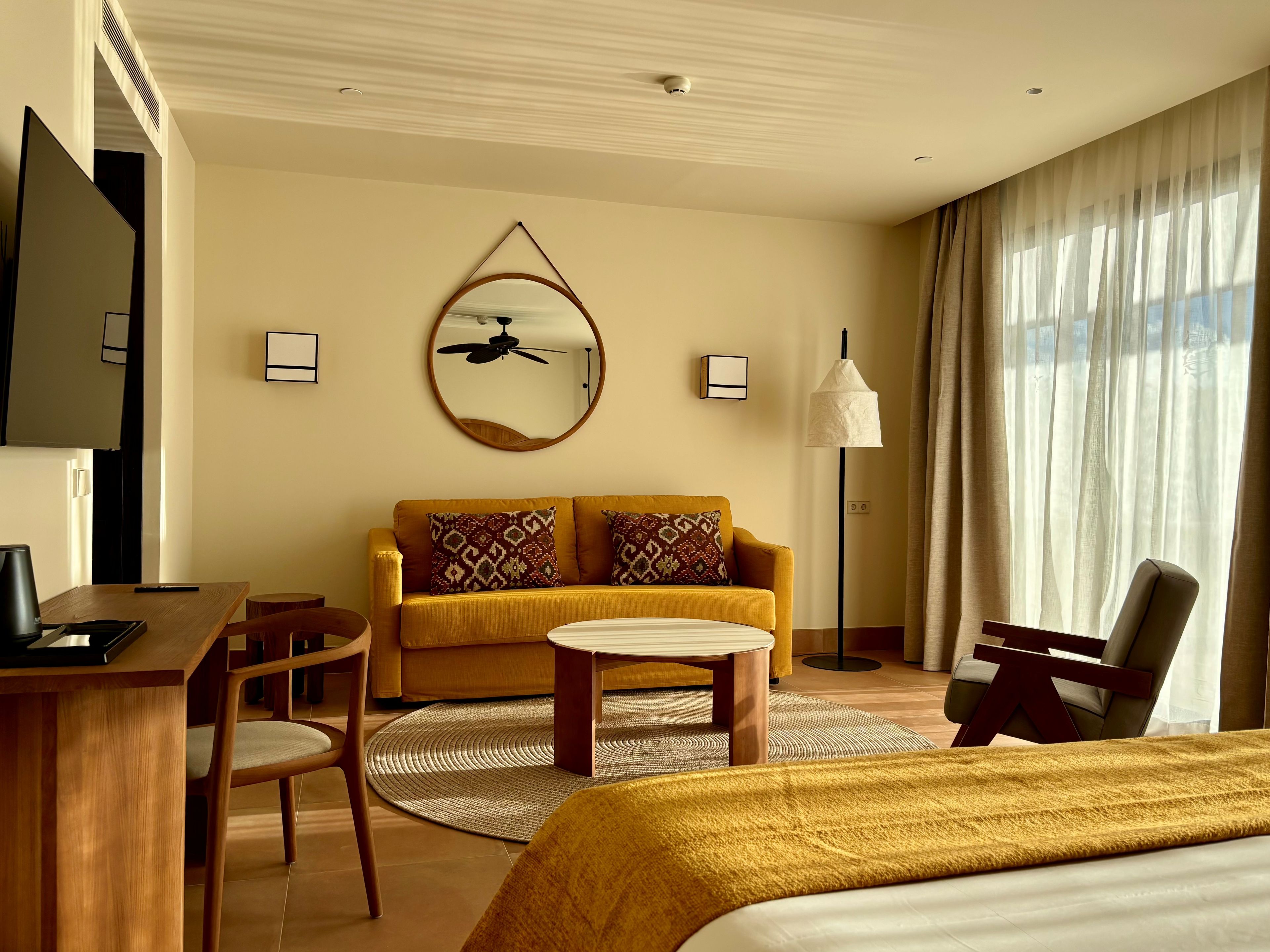 Ein Hotelzimmer mit Bett, Sofa, Schreibtisch und Sessel. Es gibt einen Deckenventilator, eine Lampe und ein an der Wand befestigtes Fernsehgerät.