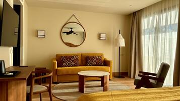 Ein Hotelzimmer mit Bett, Sofa, Schreibtisch und Sessel. Es gibt einen Deckenventilator, eine Lampe und ein an der Wand befestigtes Fernsehgerät.