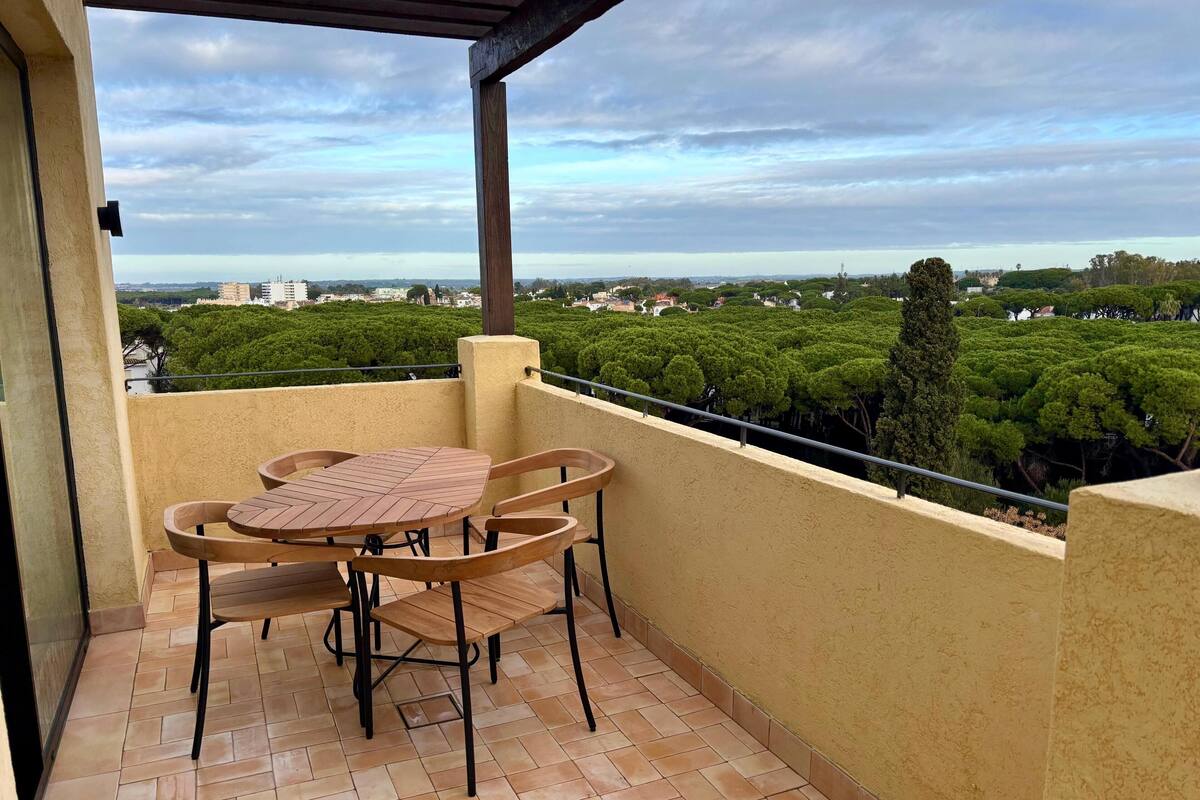 Ein Balkon mit Tisch und Stühlen, ein Fliesenboden sowie ein Blick auf eine grüne Landschaft und Gebäude.
