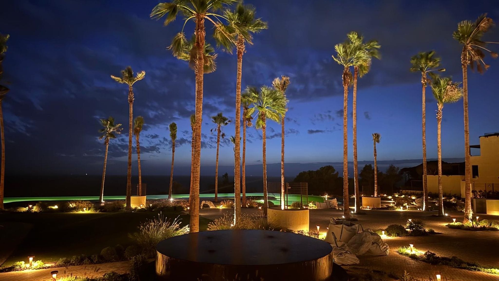 Une vue nocturne d’un complexe hôtelier avec des palmiers, une piscine et une allée bien éclairée.