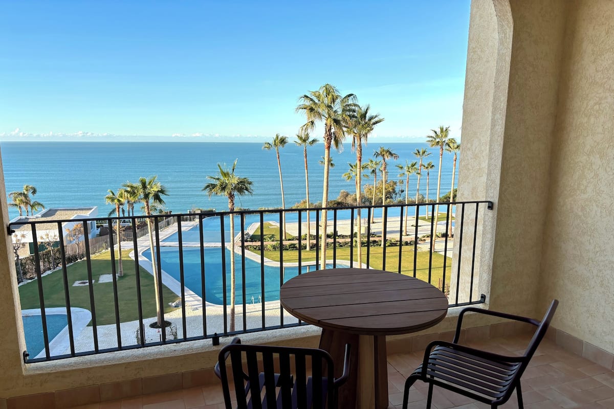 Un balcon donnant sur une piscine et des palmiers, une table ronde et deux chaises.