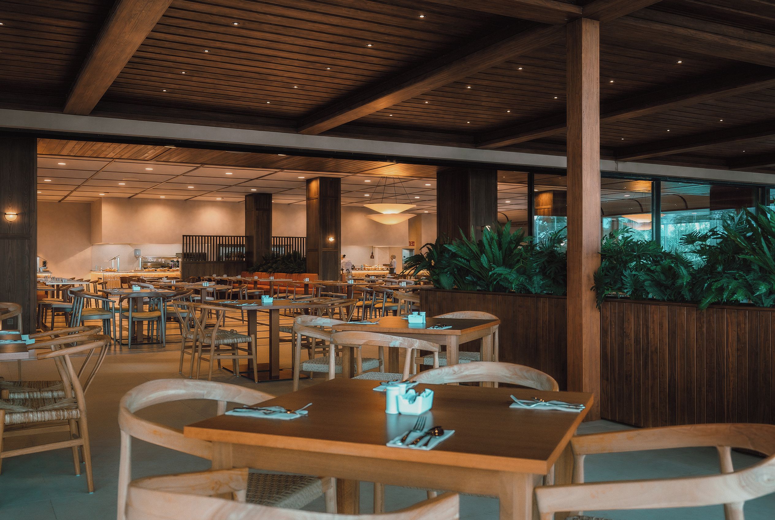 Un restaurante espacioso con mesas y sillas de madera, techo alto con vigas de madera y grandes ventanales con vistas a un jardín.