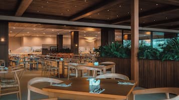 Un restaurante espacioso con mesas y sillas de madera, techo alto con vigas de madera y grandes ventanales con vistas a un jardín.