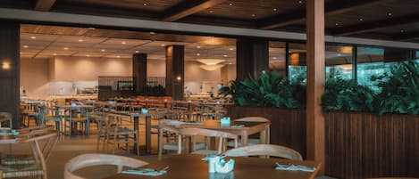 A spacious restaurant with wooden tables and chairs, a high ceiling with wooden beams, and large windows overlooking a garden.