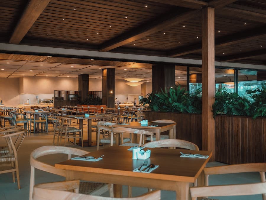 A spacious restaurant with wooden tables and chairs, a high ceiling with wooden beams, and large windows overlooking a garden.