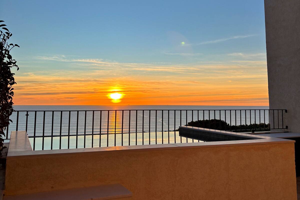 Ein Balkon mit Blick auf den Sonnenuntergang über dem Meer, inklusive Geländer und Treppe, die nach unten führt.