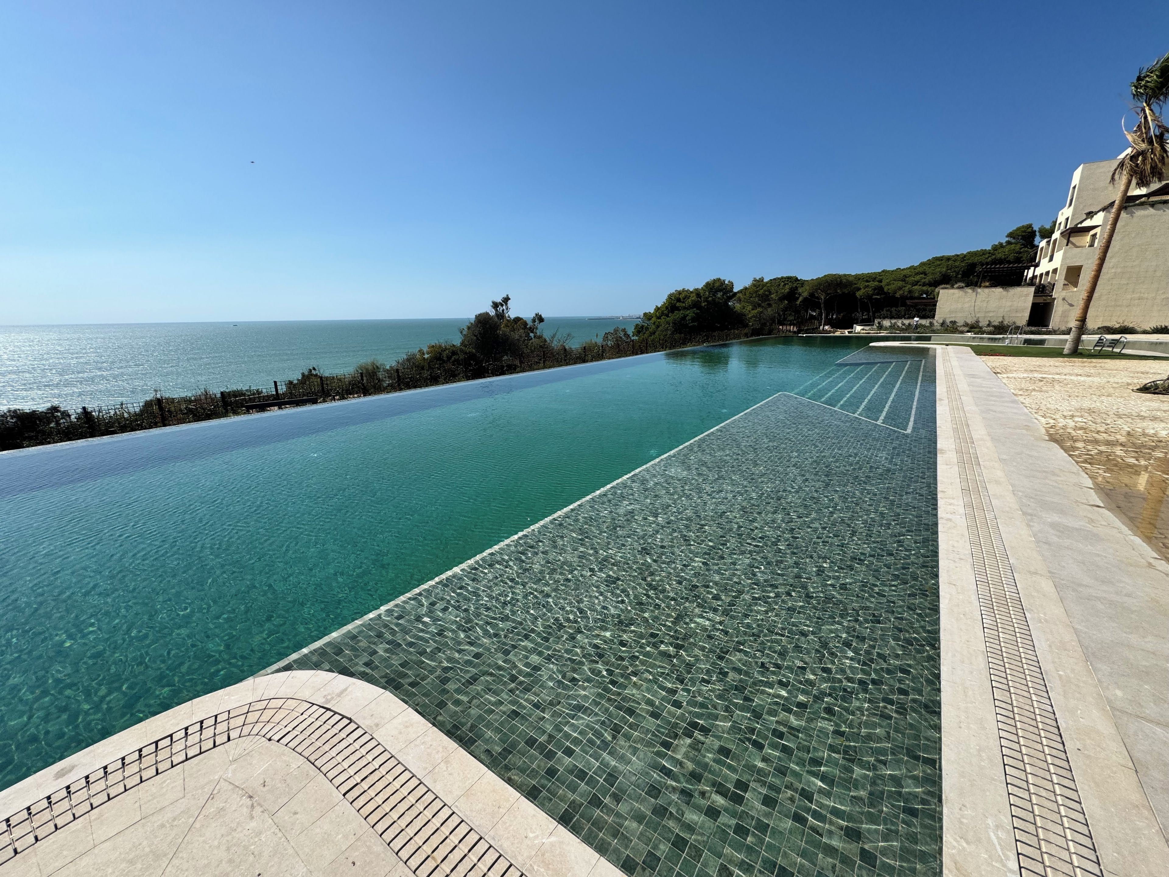 Una piscina infinita transparente con vista al mar, un camino empedrado y un edificio con una palmera.