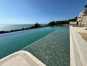 A clear infinity pool with a view of the sea, a tiled walkway, and a building with a palm tree. - Punta Bermeja Premium Beach Hotel (El Puerto de Santa Maria)