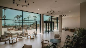 Un espacio interior amplio con ventanas grandes, una zona de estar con sofás y mesas, y vistas a palmeras y un cuerpo de agua.