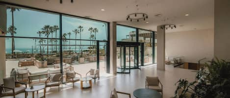 A spacious indoor area with large windows, a seating area with sofas and tables, and a view of palm trees and a body of water.