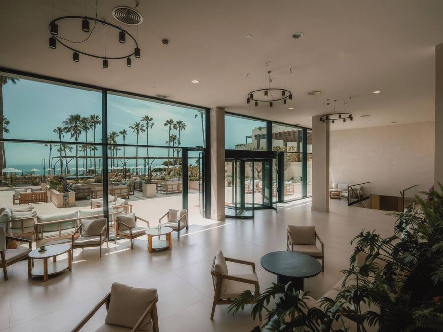 Un espacio interior amplio con ventanas grandes, una zona de estar con sofás y mesas, y vistas a palmeras y un cuerpo de agua.