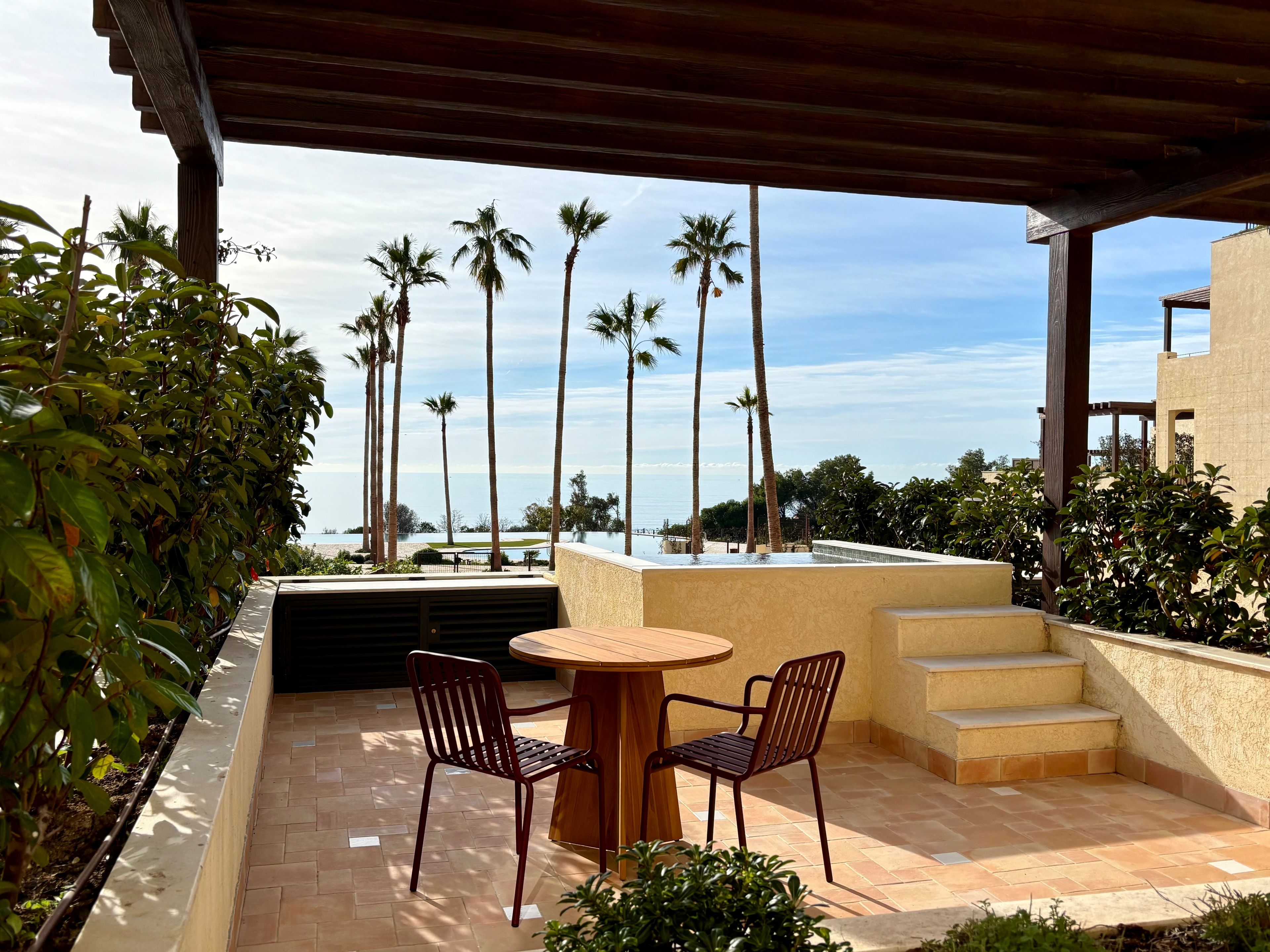 Ein überdachter Außenbereich mit Tisch und Stühlen, mit Blick auf einen Pool und Palmen.