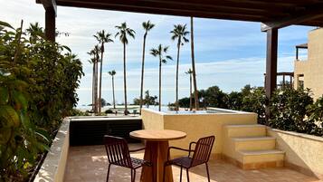 Ein überdachter Außenbereich mit Tisch und Stühlen, mit Blick auf einen Pool und Palmen.