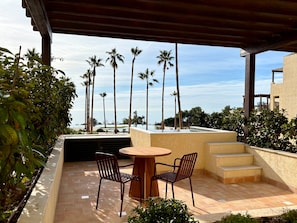 A covered outdoor seating area with a table and chairs, overlooking a pool and palm trees. - Punta Bermeja Premium Beach Hotel (El Puerto de Santa Maria)