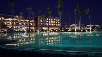 Een nachtzicht van een hotel met een zwembad, palmbomen en verlichte balkons.
