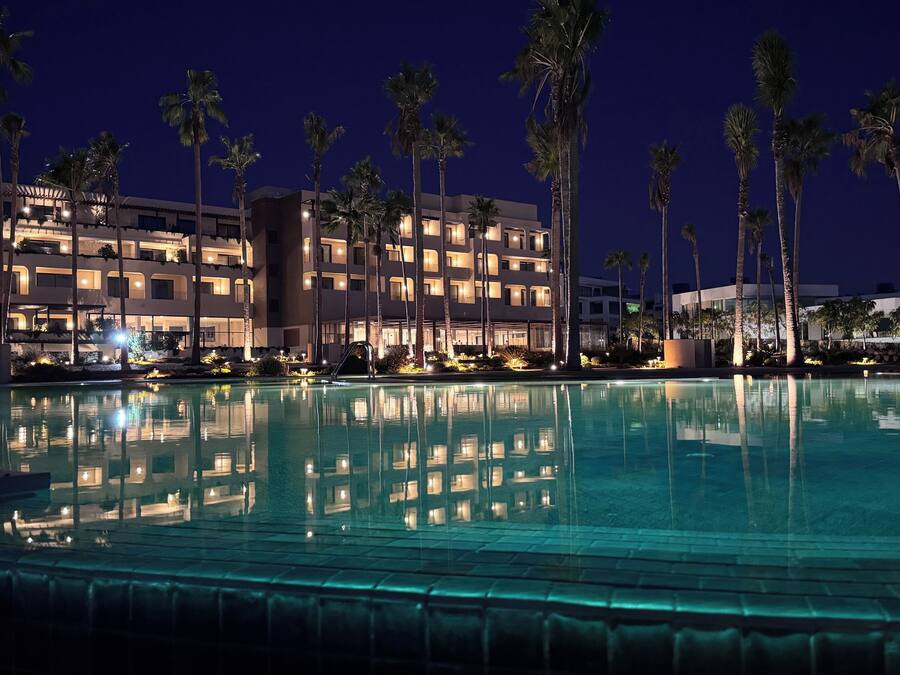 A nighttime view of a hotel with a swimming pool, palm trees, and illuminated balconies.