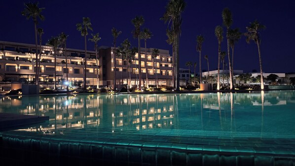 Vista nocturna de un hotel con piscina, palmeras y balcones iluminados.