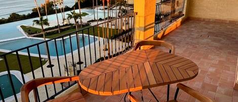 Un balcón con mesa y sillas de madera, con vistas a una piscina y palmeras.