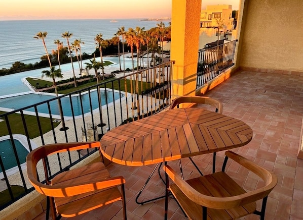 A balcony with a wooden table and chairs, overlooking a pool and palm trees. - Punta Bermeja Premium Beach Hotel (El Puerto de Santa Maria)