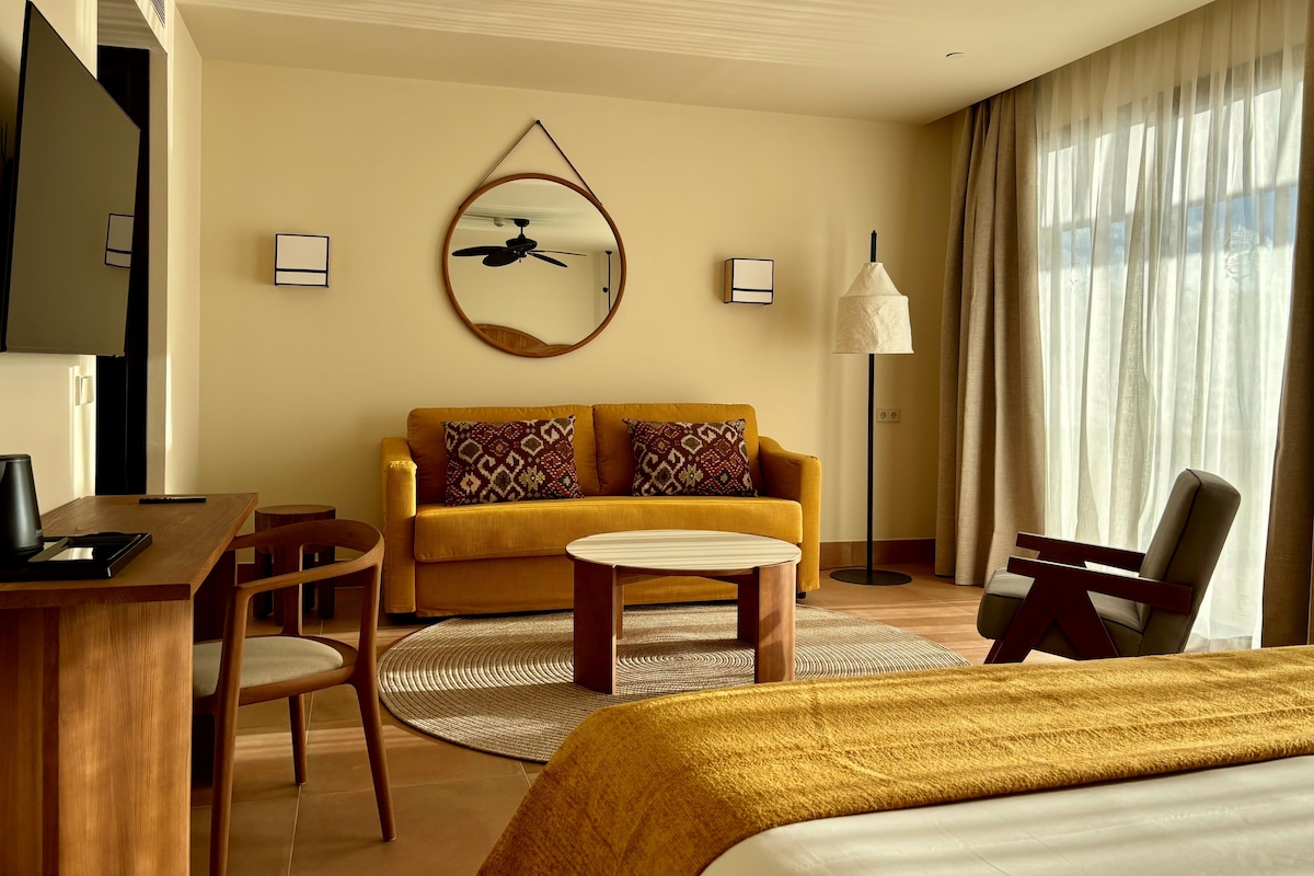 Ein Hotelzimmer mit Bett, Sofa, Schreibtisch und Sessel. Es gibt einen Deckenventilator, eine Lampe und ein an der Wand befestigtes Fernsehgerät.