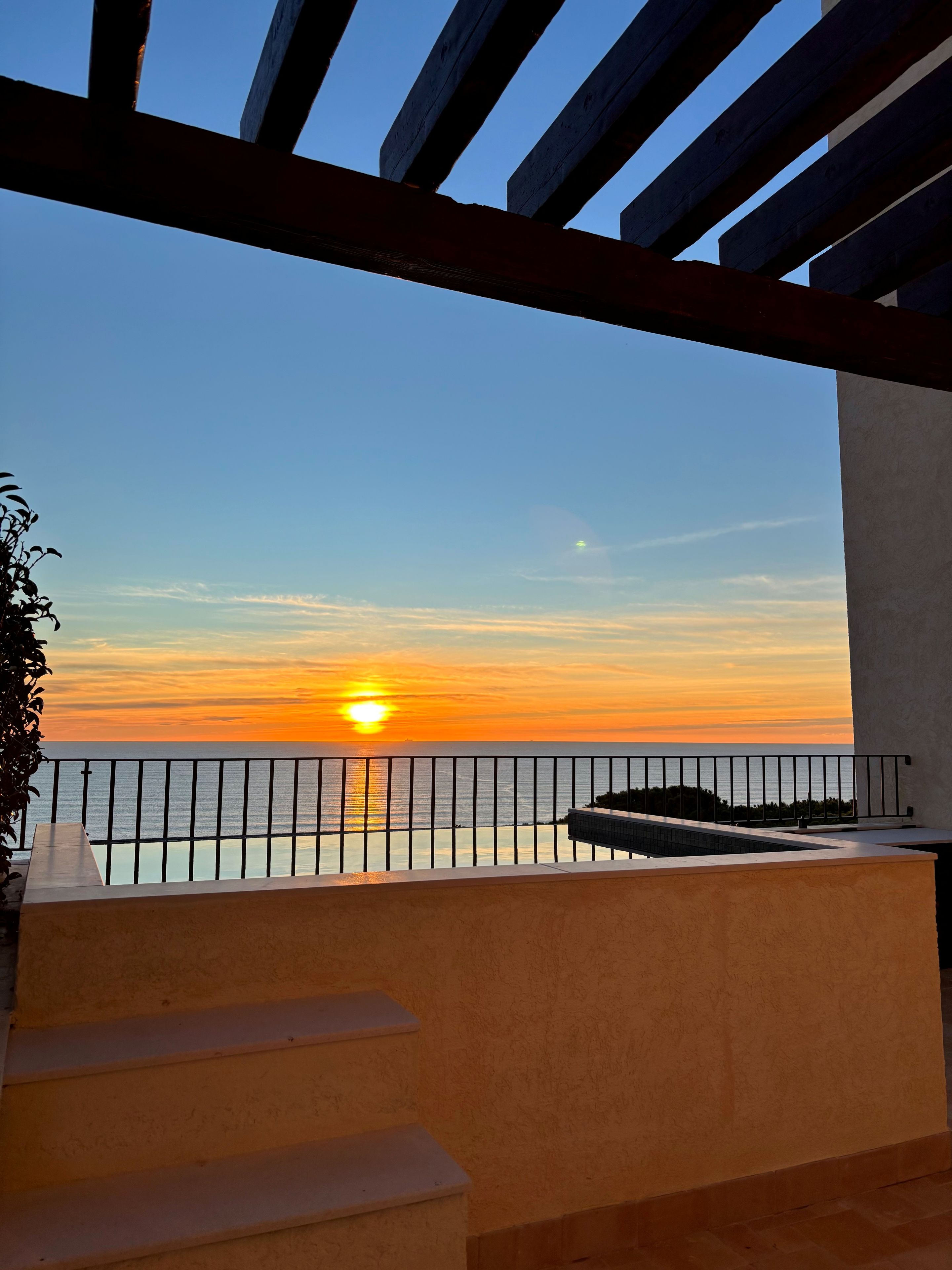 Un balcón con vistas a la puesta de sol sobre el mar, una barandilla y escaleras que bajan.