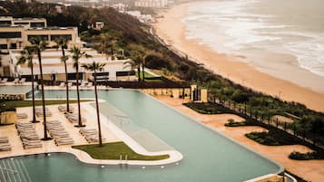 Una piscina de diseño curvo, rodeada de palmeras y sillas de descanso, contigua a una playa con arena y olas del océano.
