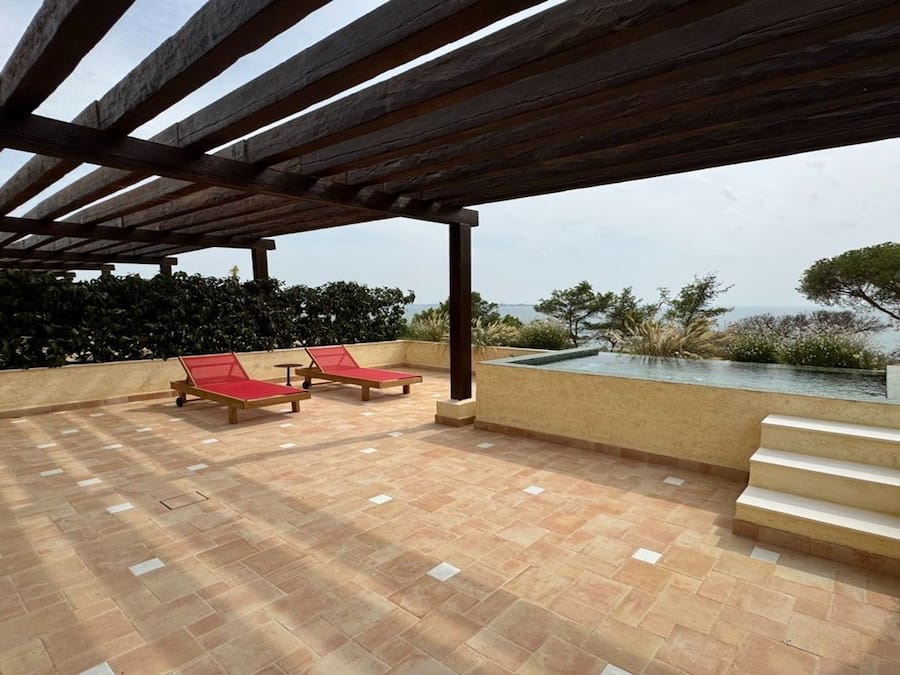 A covered outdoor area with a pool, two red lounge chairs, and a view of the sea.