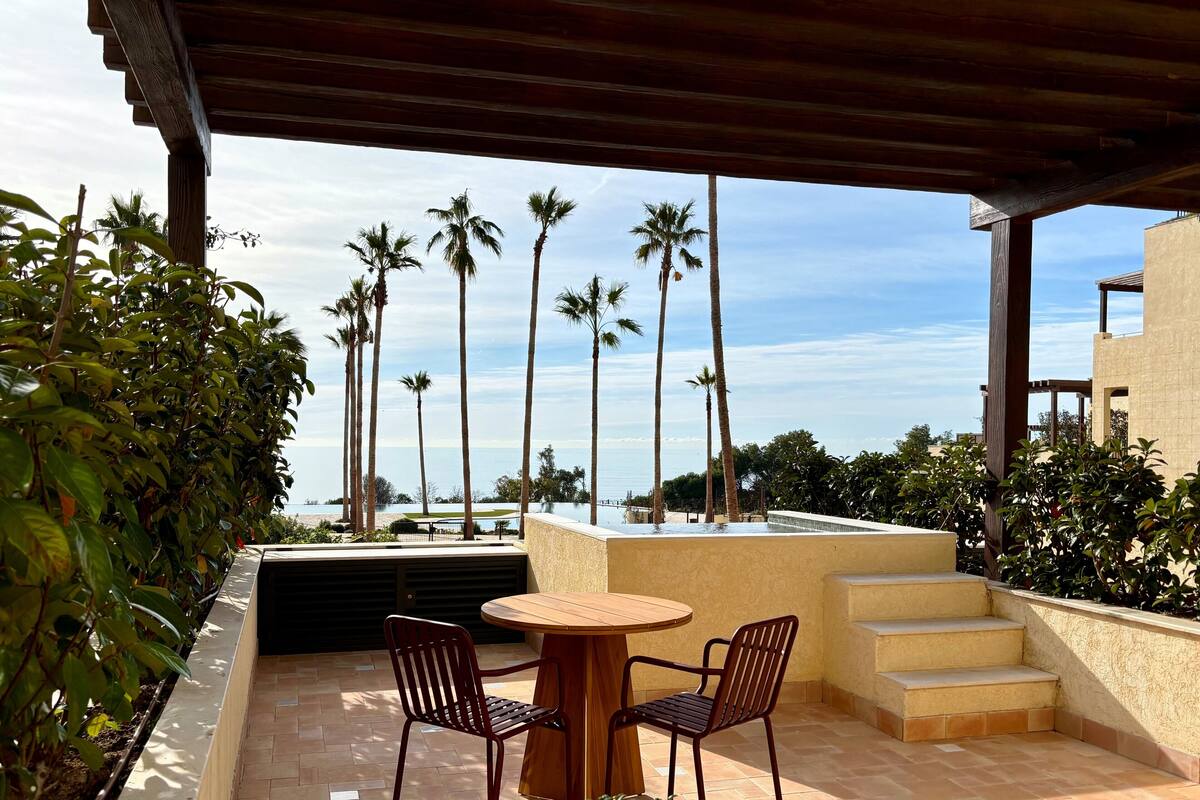 Ein überdachter Außenbereich mit Tisch und Stühlen, mit Blick auf einen Pool und Palmen.
