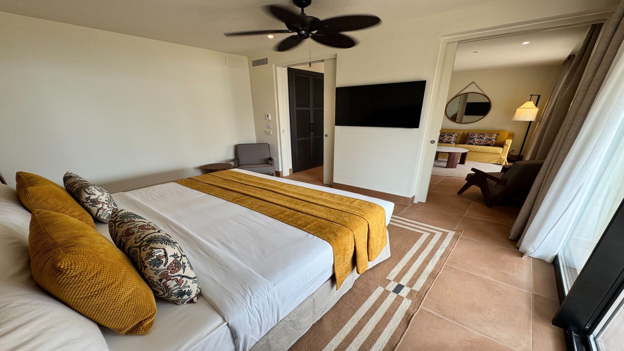 Camera d'albergo con un letto grande, una ventola a soffitto, una TV e un divano nella zona soggiorno adiacente.