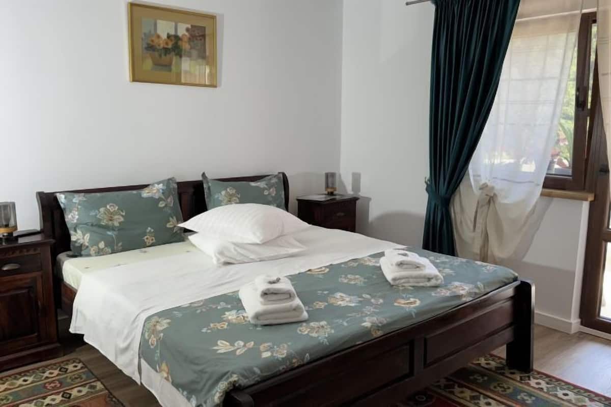 A neatly made bed with floral bedding, a bedside table with a lamp, and a window with curtains.