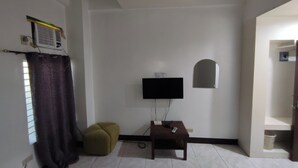 Ein kleines, schlichtes Zimmer mit einem an der Wand montierten Flachbildfernseher, einem kleinen Tisch mit Fernbedienung, einem Wandheizkörper und einem Fenster mit Vorhang.