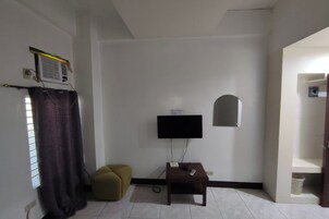 A small, simple room with a flat-screen TV mounted on the wall, a small table with a remote control, a heater on the wall, and a window with a curtain.