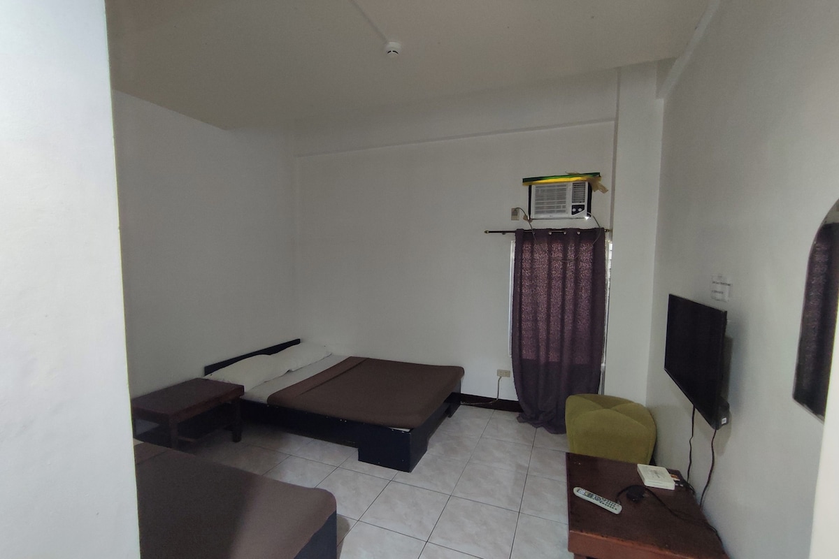 Una habitación pequeña y poco amoblada con una cama, un escritorio, una silla, un televisor y una ventana con cortinas.