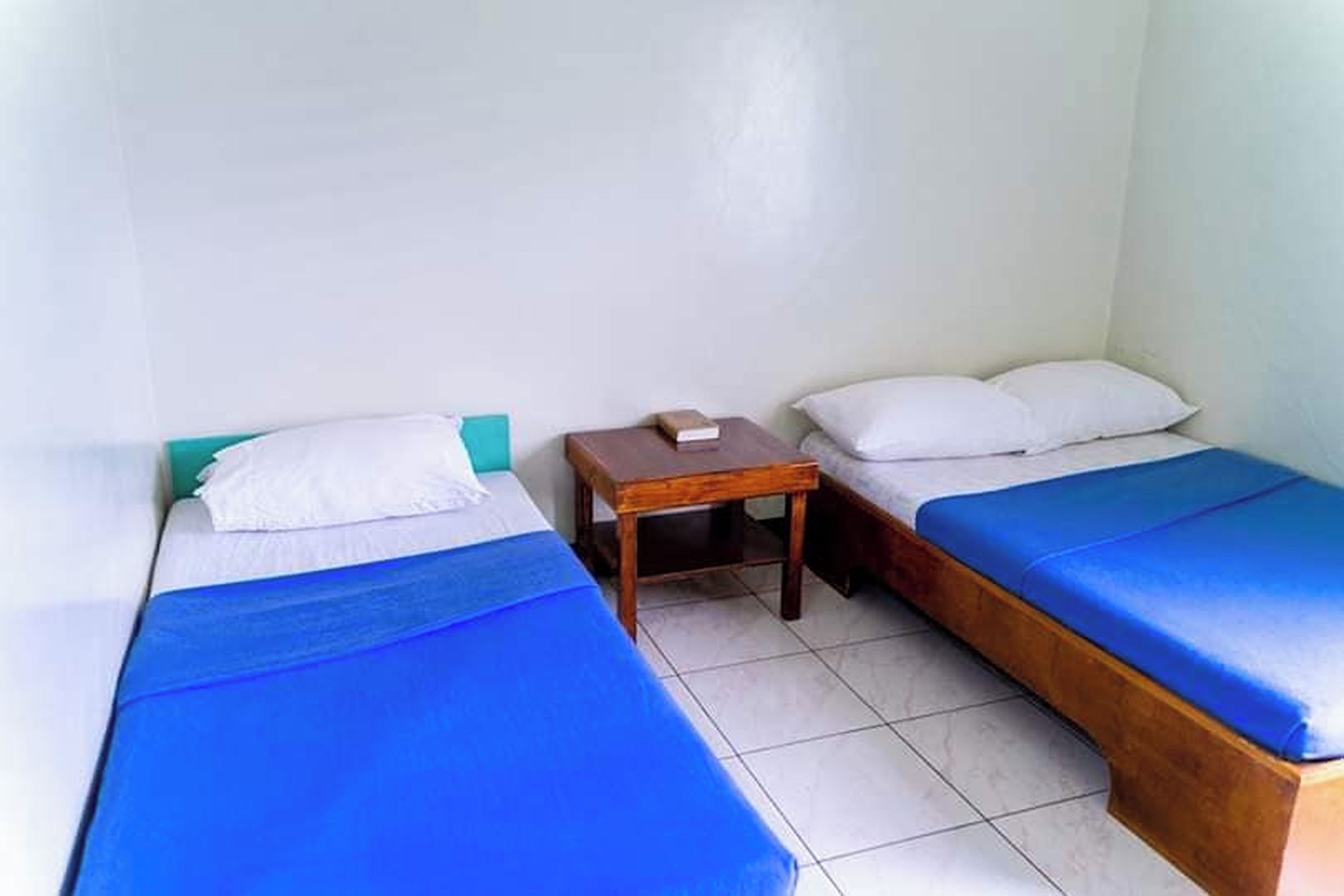 A small, clean room with two single beds, each with a blue bedspread and white pillows. There is a wooden nightstand between the beds.
