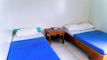 A small, clean room with two single beds, each with a blue bedspread and white pillows. There is a wooden nightstand between the beds.