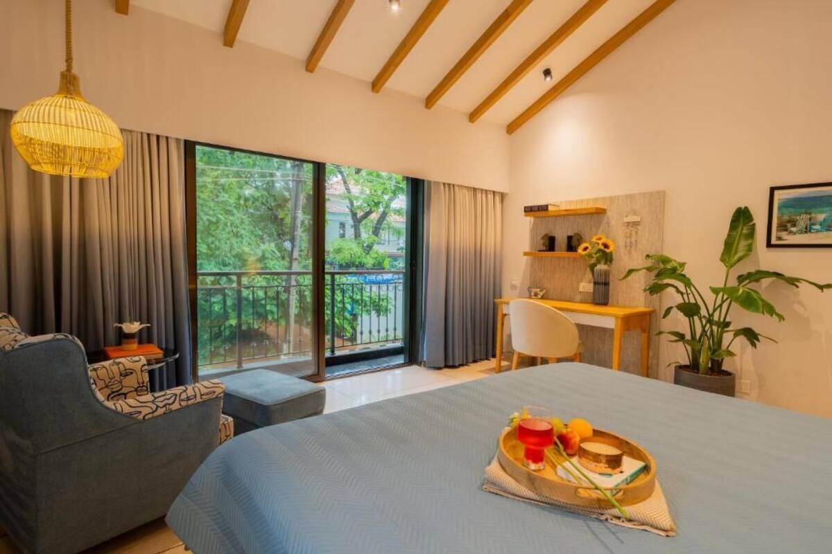 A bedroom with a bed, a desk, a chair, a hanging light fixture, and a view of greenery through the window.