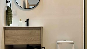 A modern bathroom with a wooden vanity, a round mirror, a toilet, and a towel rack. - Makihi Suites (Hanga Roa)