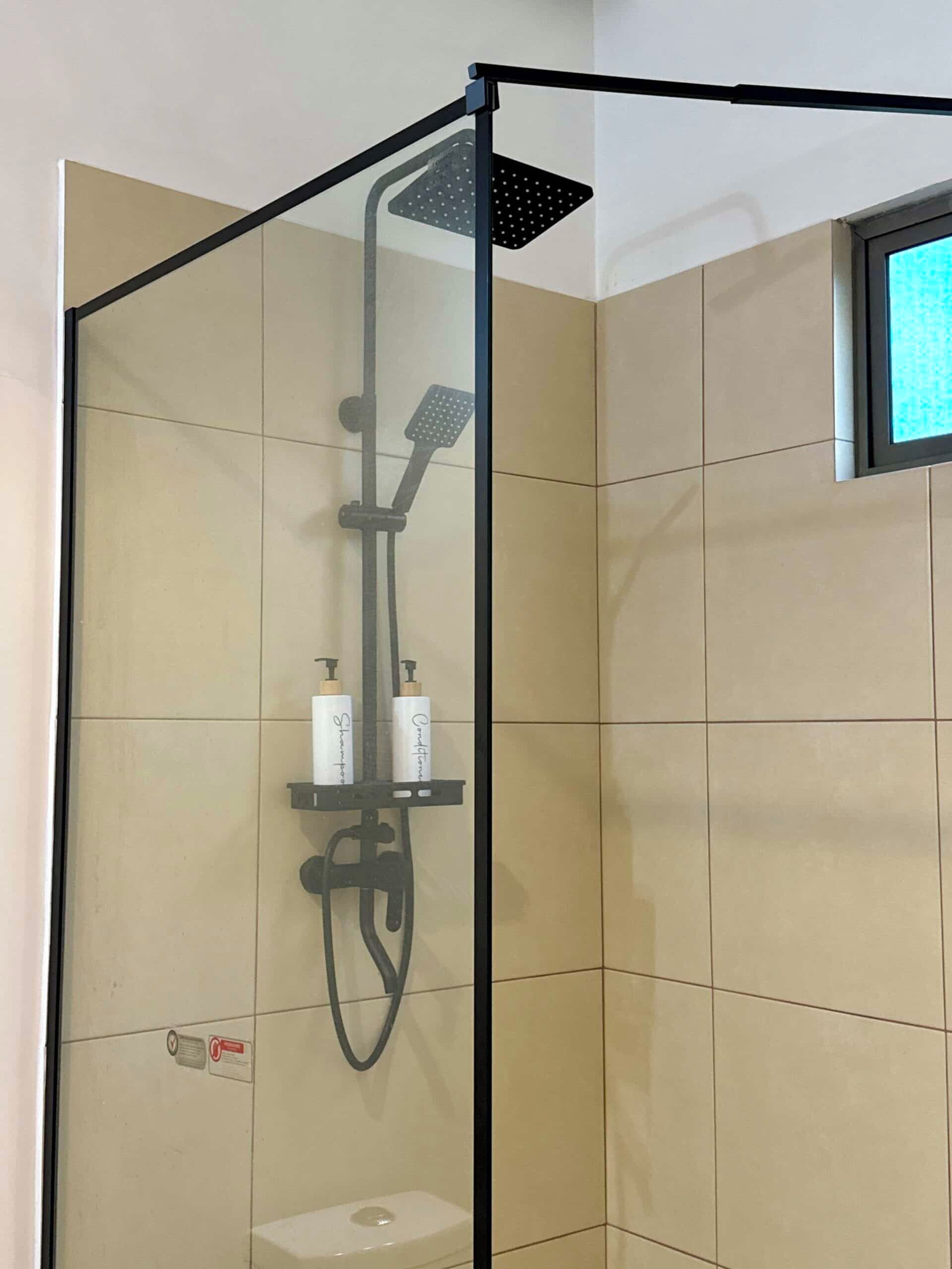 A modern shower enclosure with a handheld showerhead, a rain showerhead, and a handheld scrubber. There are two bottles of shampoo and conditioner on a shelf.
