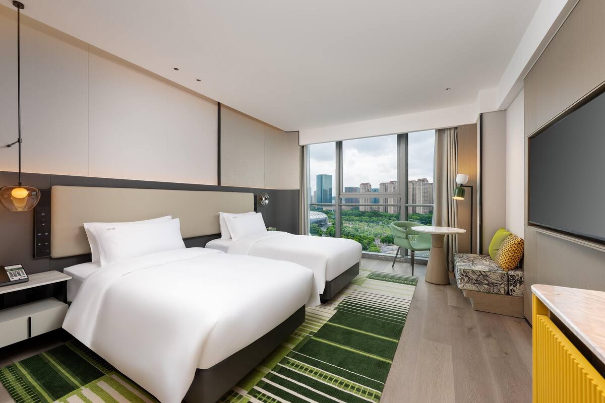 A modern hotel room with two beds, a desk, a chair, and a large window offering a city view.